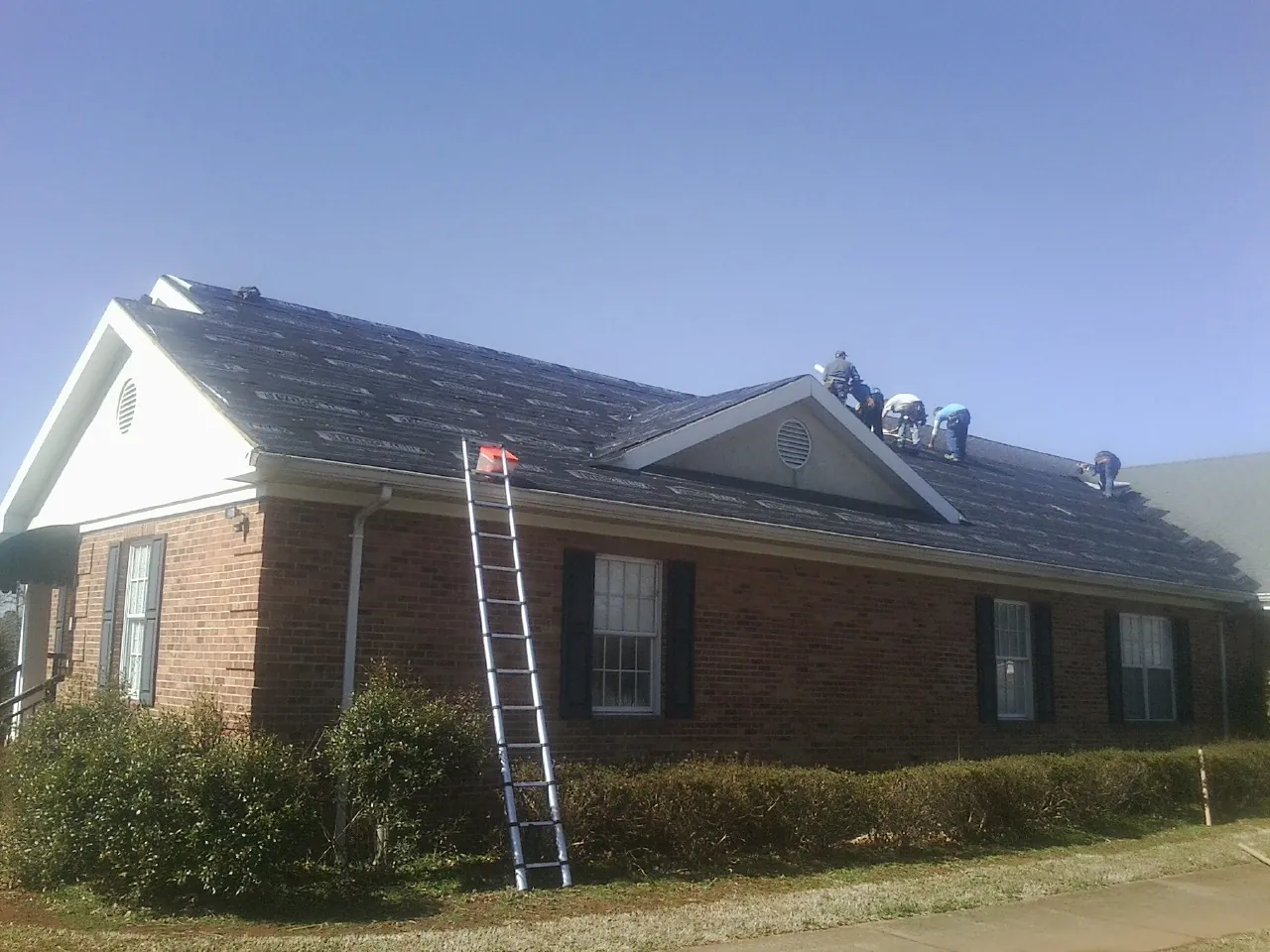 Crew tearing shingles off a brick ranch roof with a ladder set — Emergency Roof Repair work by our crew in Edgewood